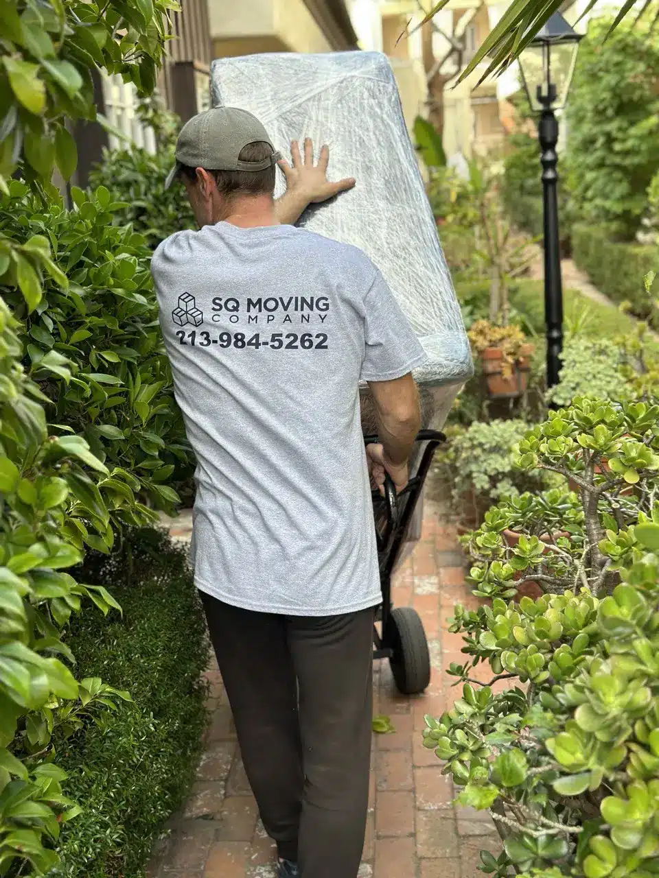 Mover transporting a shrink-wrapped item on a hand truck through a narrow walkway during a Long Beach move.
