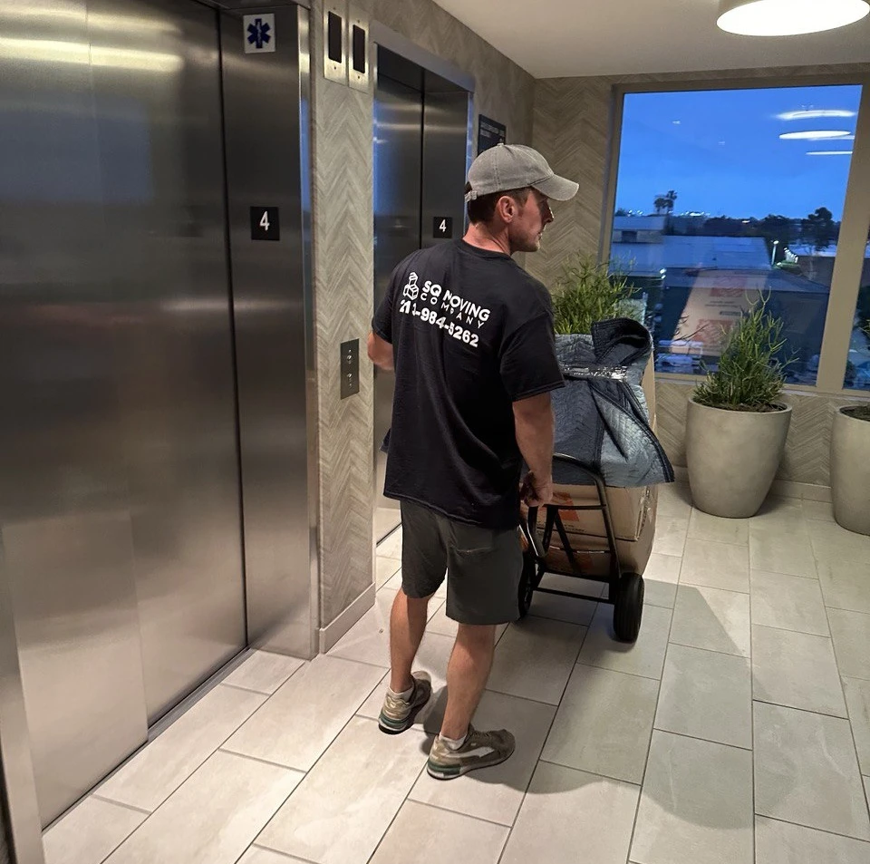 SQ Moving Company mover pushing a padded load on a dolly toward an elevator in a modern apartment building in Century City