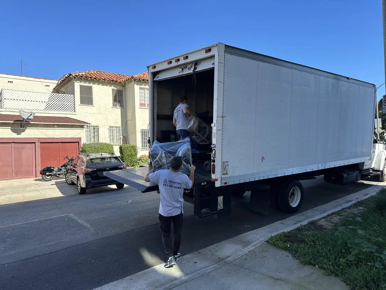 SQ Moving Company crew loading wrapped furniture into a large moving truck on a residential Los Angeles street under a clear blue sky